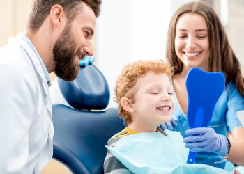 Young boy looking at the mirror with toothy smile sitting on the chair with dentist and assistant at the dental office