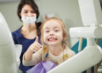 Cheerful kid with broad smile after painless teeth polishing procedure sits in comfortable chair at modern dentist office with mother and doctor in ask who stand behind.